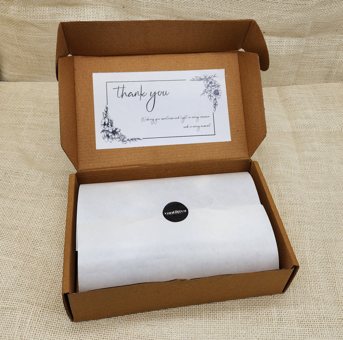 Open cardboard box with a white sheet of paper and a 'thank you' card on a beige fabric background

